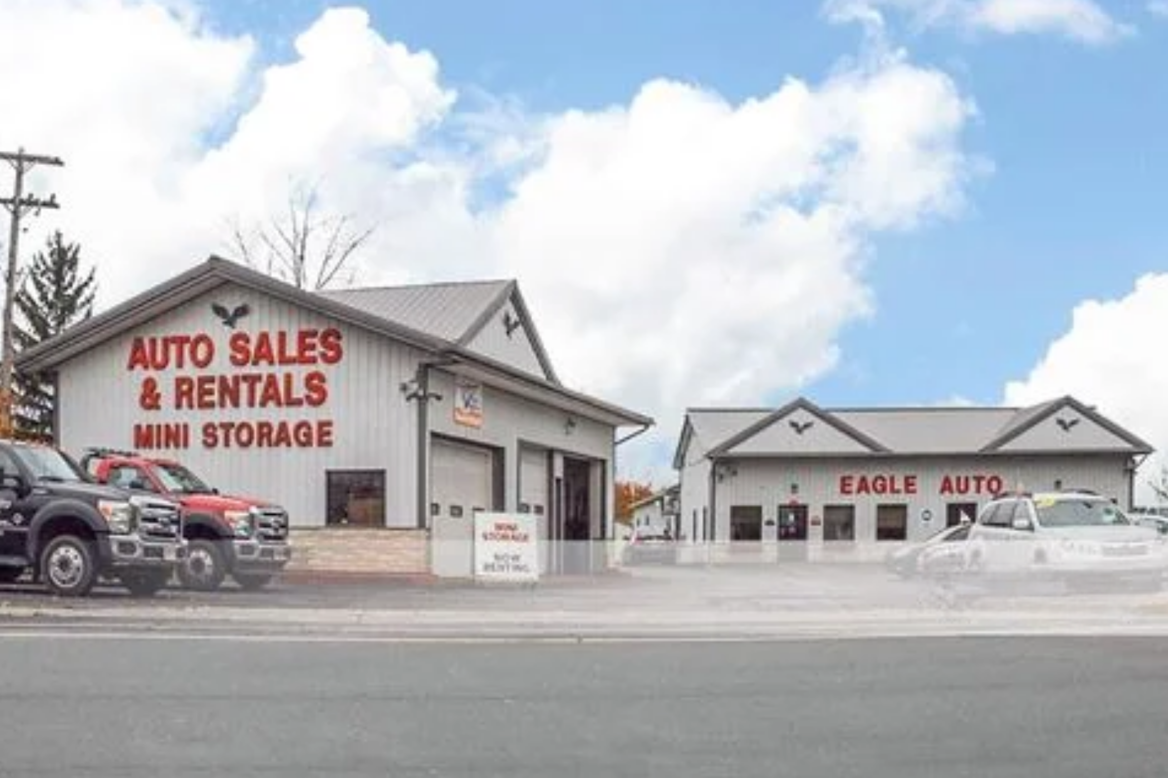 Auto sales and rental business with gray metal buildings labeled “Auto Sales & Rentals Mini Storage” and “Eagle Auto.” Several trucks and cars are parked in front under a partly cloudy sky.